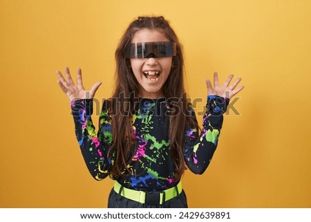 Adorable hispanic girl, mad with joy, celebrates excited virtual reality win, arms raised, glasses on. triumph personified over yellow isolated background