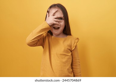 Adorable hispanic girl standing peeking in shock covering face and eyes with hand, looking through fingers afraid over isolated yellow background - Powered by Shutterstock