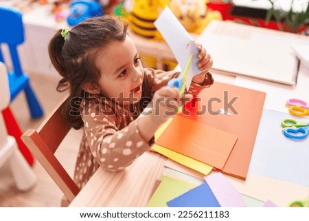 Adorable hispanic girl student smiling confident cutting paper at kindergarten
