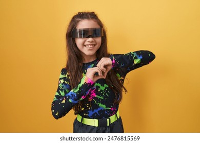 Adorable hispanic girl in virtual reality glasses forms heart symbol with hands, radiating love on isolated yellow background - Powered by Shutterstock