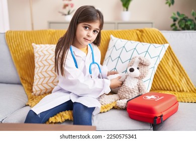 Adorable hispanic girl wearing doctor uniform bandaging teddy bear arm at home - Powered by Shutterstock