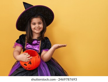 Adorable hispanic girl in witch costume, confidently celebrating her halloween win with a jubilant smile, while holding a pumpkin basket on an isolated yellow background - Powered by Shutterstock