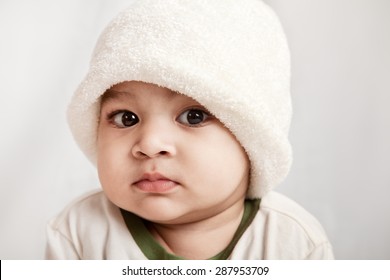 Adorable Indian Baby Boy In White Cap Looking At Camera Over Gray Background