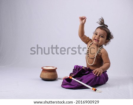 adorable Indian baby in krishna kanha or kanhaiya dress posing with his flute and dahi handi (pot with curd) on white background. sitting pose