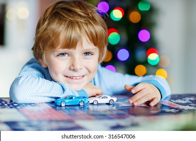 Adorable Kid Boy Playing With Cars And Toys At Home, Indoor. Funny Child Having Fun With Gifts. Colorful Christmas Lights On Background. Family, Holiday, Kids Lifestyle Concept.