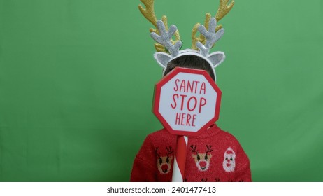 Adorable kid with double headband christmas deer antlers showing sign santa stop here. Isolated on green. High quality photo - Powered by Shutterstock