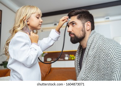 Adorable Kid In Medical Lab Coat Pressing Stethoscope To Forehead Of Her Father Stock Photo