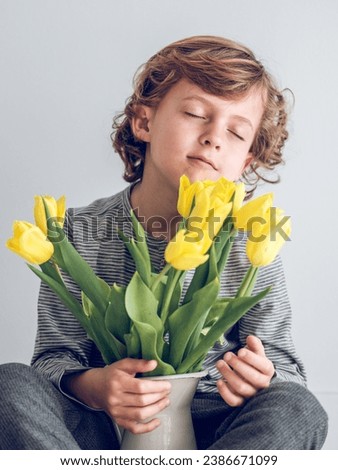 Adorable kid smelling bunch of yellow tulip flowers in vase and enjoying fresh scent with closed eyes on white background