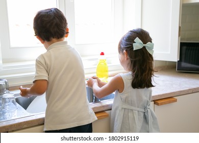 Adorable Kids Washing Plates And Glasses At Kitchen. Back View Of Children Helping Parents And Cleaning Dishes After Breakfast. Housekeeping And Family Concept