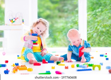 Adorable laughing toddler girl and a funny little baby boy, brother and sister, playing with colorful blocks sitting on a floor in a sunny bedroom with a big window 