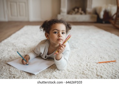 Adorable Little African American Girl Drawing With Pencils And Lying On Carpet At Home