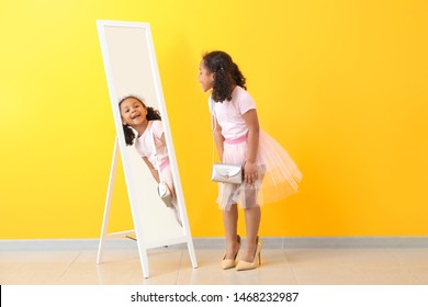 Adorable little African-American girl with mother's shoes looking into mirror near color wall