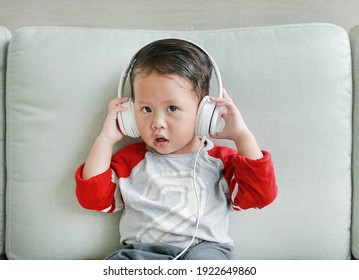 Adorable little Asian baby boy in headphones is using a smartphone lying on the sofa at home. Child listening to music on earphones.