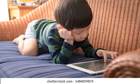 Adorable Little Asian Boy Lying On Sofa Alone Playing Tablet Computer. Digital Addiction, Screen Time, Gamification, Fun, E-learning, Technology And Kids, Modern Lifestyle, Child Development.