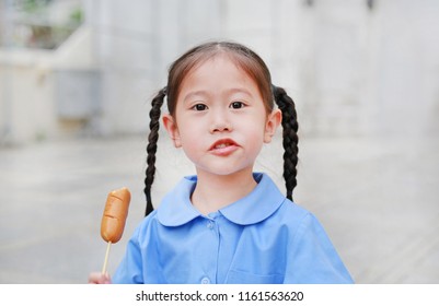 Adorable little Asian child girl in school uniform enjoy eating sausage.
