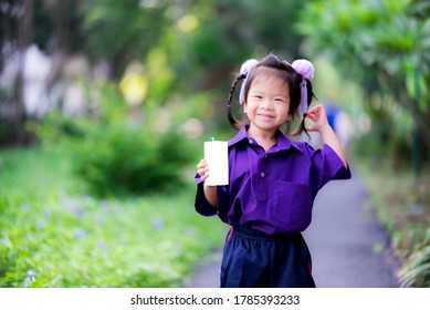 Adorable Little Asian Child Girl Holding White Milk Box And Drink On Green Natural Background. Kid Wearing Purple School Uniform And Sweet Smiling In The Morning. Children Aged 3 - 4 Years Old.