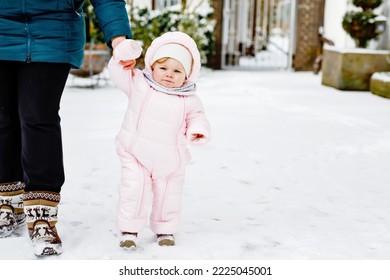 Adorable Little Baby Girl Making First Steps Outdoors In Winter Through Snow. Cute Toddler Learning Walking. Mother Holding Child On Hand. Daughter And Mum Walk Together. Happy Family