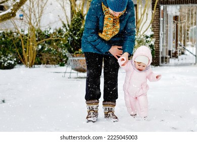 Adorable Little Baby Girl Making First Steps Outdoors In Winter Through Snow. Cute Toddler Learning Walking. Mother Holding Child On Hand. Daughter And Mum Walk Together. Happy Family