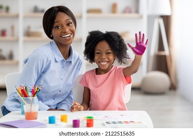 Adorable Little Black Girl With Curly Hair Having Fun At Daycare, Happy Kid Painting Under Child Development Specialist Attractive Young Woman, Showing Palm Colored With Paints And Smiling