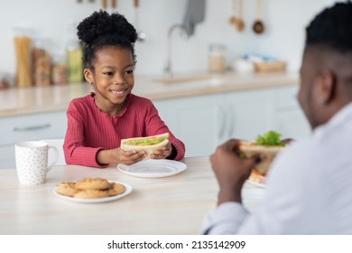 Adorable Little Black Girl Looking At Her Dad While Eating Sandwich, Happy Black Father And Kid Sitting In Front Of Each Other At Kitchen Table, Having Healthy Breakfast Together At Home