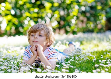 Adorable little blond child with blue eyes laying on the grass with daisies flowers in the park. On warm summer day during school holidays. Kid boy dreaming and smiling.