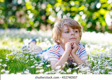 Adorable little blond child with blue eyes laying on the grass with daisies flowers in the park. On warm summer day during school holidays. Kid boy dreaming and smiling.