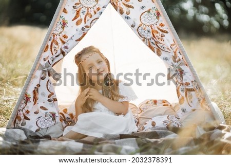 Adorable little blond girl sitting in boho teepee tent. Cute girl child, wearing white dress and feather hair accessories in natice american style, posing to camera in wigwam outdoors in summer field