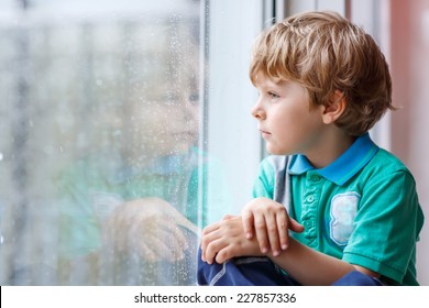 Adorable Little Blond Kid Boy Sitting Near Window And Looking On Raindrops, Indoors.