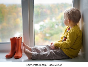 Adorable Little Blond Kid Boy Sitting On The Windowsill With Rain Shoes And Looking On Autumn Raindrops, Indoors. A Child Looks Out The Window. Vintage Color.  Orange Rubber Boots Standing Near Window