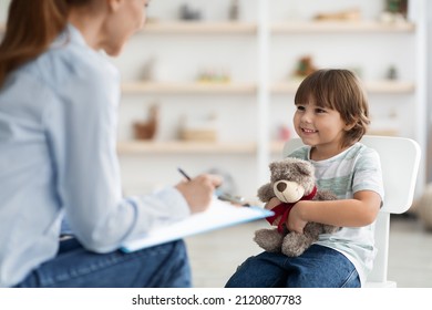 Adorable little boy hugging teddy bear and smiling to specialist sitting at psychotherapist office, professional kids psychologist talking to small patient - Shutterstock ID 2120807783