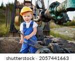 Adorable little boy looking at camera and smiling while tightening bolt on pipe in oil field. Cute kid in construction helmet using industrial wrench while repairing pump jack pipe outdoors.