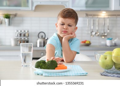 Adorable little boy refusing to eat vegetables at table in kitchen