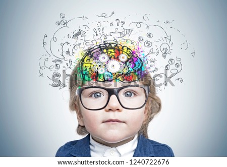 Adorable little boy wearing blue suit and glasses standing near gray wall with colorful brain sketch with cogs drawn inside his head. Concept of thinking