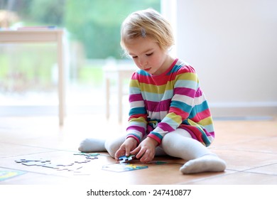 Adorable little child, blonde toddler girl, playing with puzzles sitting on the tiles floor next to a big window at home or kindergarten