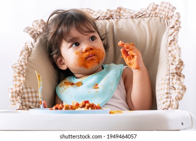 Adorable Little Child Funny Girl Eating Spaghetti With Spoon While Sitting In High-powered Chair At Home. Toddler Child With Tomato Sauce Making Mess Her Face Looking At Parent. Self-feeding Concept