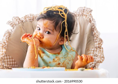 Adorable little child funny girl eating spaghetti with spoon while sitting in high-powered chair at home. Toddler child with tomato sauce making mess her face looking at parent. Self-feeding concept