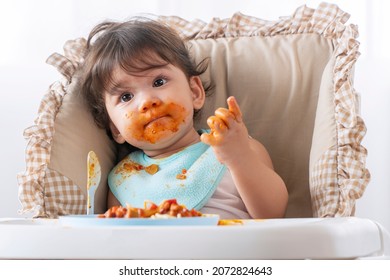 Adorable Little Child Funny Girl Eating Spaghetti With Spoon While Sitting In High-powered Chair At Home. Toddler Child With Tomato Sauce Making Mess Her Face Looking At Parent. Self-feeding Concept