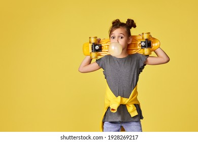 Adorable Little Child Girl With Yellow Skateboard Blowing A Big Bubble Gum Over Yellow Background. Copy Space