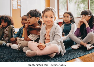 Adorable little child looking at the camera while sitting in class with other kids. Girl attending a lesson in elementary school. Primary education and early child development.