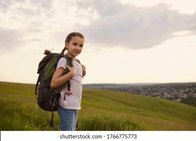Adorable little girl with backpack standing on mountain top, copy space. Cute kid with rucksack hiking in countryside
