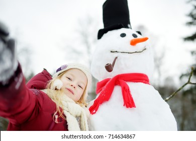 Adorable Little Girl Building A Snowman In Beautiful Winter Park. Cute Child Playing In A Snow. Winter Activities For Kids.