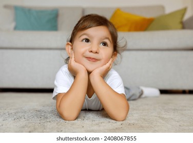 Adorable little girl with a charming smile lies on her stomach on the floor, hands on cheeks, cute toddler female child looking up with a dreamy, joyful expression in cozy home setting, copy space - Powered by Shutterstock