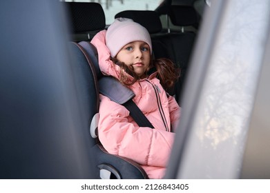 Adorable Little Girl, Cheerful Child In Pink Warm Clothes Sitting Fastened With A Secure Seat Belt In The Modern Car During A Family Winter Trip By Car. Concept Of A Safe Travel With Kids