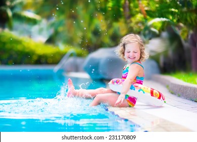 Adorable Little Girl With Curly Hair Wearing A Colorful Swimming Suit Playing With Water Splashes At Beautiful Pool In A Tropical Resort Having Fun During Family Summer Vacation