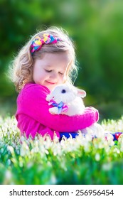 Adorable Little Girl, Cute Curly Toddler In A Colorful Summer Dress, Playing With A Real Rabbit, Having Fun With Her Pet Bunny In A Beautiful Garden With First Spring Snowdrop Flowers