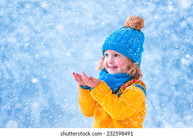 Adorable little girl, cute toddler in a blue knitted hat and yellow Nordic sweater, playing with snow catching snowflakes having fun outdoors in a beautiful winter park