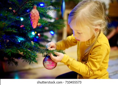 Adorable Little Girl Decorating The Christmas Tree With Colorful Glass Baubles. Trimming The Christmas Tree. Celebrating Xmas At Home.