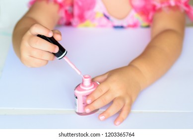 Adorable Little Girl Having Fun Playing At Home With Colorful Pink Nail Polish Doing Manicure And Painting Nails. Toddler Cute  Child In Pink Elegant Dress Sitting Wood White Table And Making Manicure