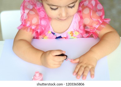 Adorable Little Girl Having Fun Playing At Home With Colorful Pink Nail Polish Doing Manicure And Painting Nails. Toddler Cute  Child In Pink Elegant Dress Sitting Wood White Table And Making Manicure