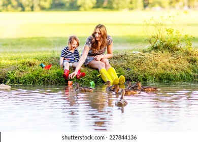 Adorable Little Girl And Her Mom Playing With Paper Boats And Feeding Ducks In A River. Creative Leisure With Kids.